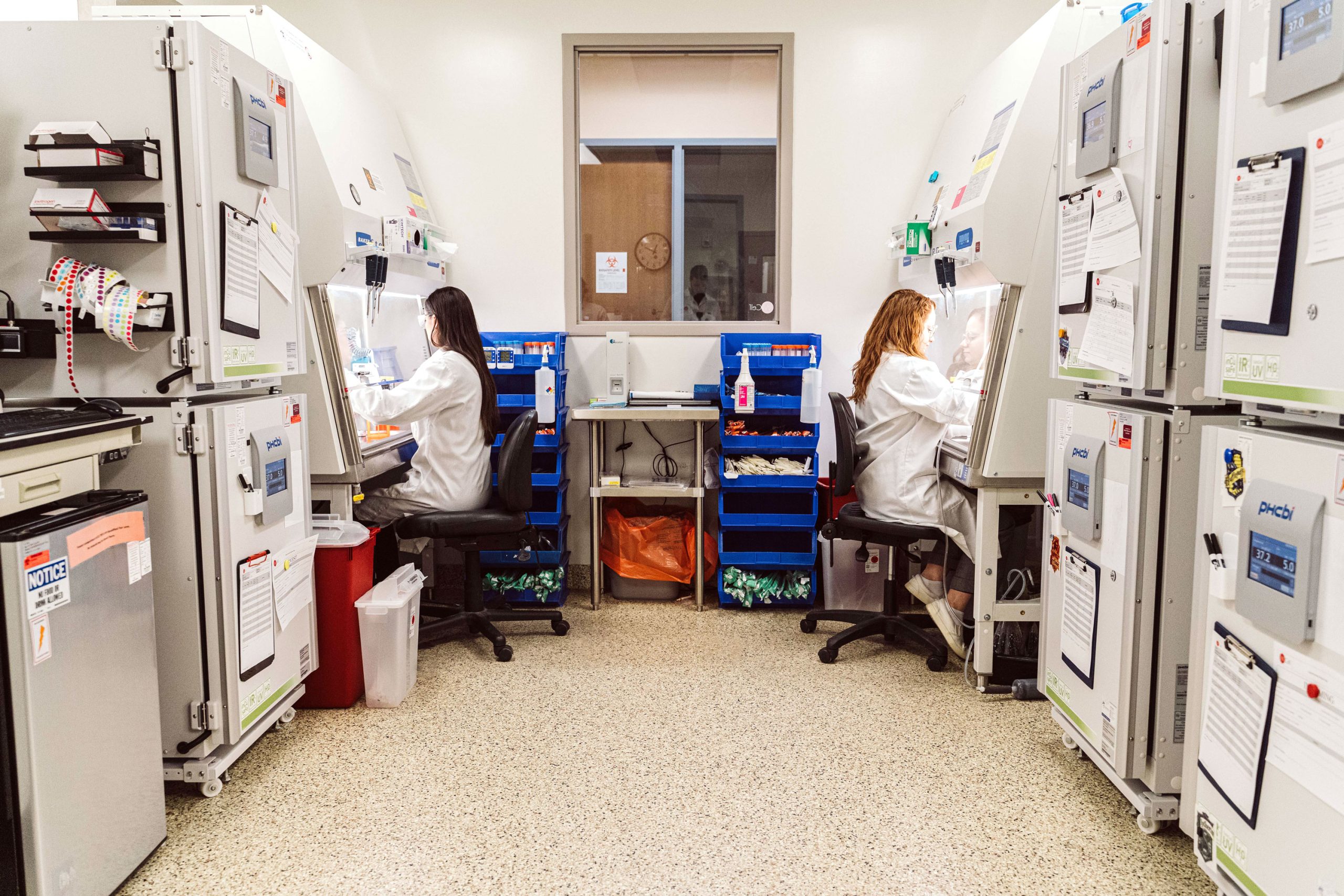 Two scientists sit at fume hoods on opposite sides of a laboratory.